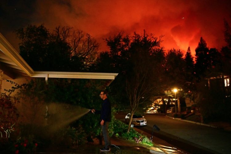 A man waters the front of his house as smoke and flames from the Palisades Fire burn toward the Encino neighborhood of Los Angeles. ©AFP